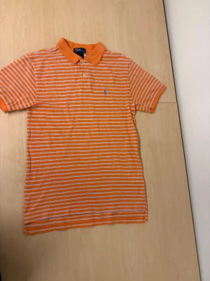 Randig orange piké från Ralph Lauren - Snygg orange pikétröja från Ralph Lauren med vita horisontella ränder och klassisk krage. Tröjan har korta ärmar, knäppning framtill och den ikoniska blå loggan broderad på bröstet. Perfekt för dig som gillar stilrena och sportiga plagg.