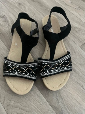 Svarta sandaler från Gyllene Gripen - Snygga svarta sandaler från Gyllene Gripen med bred rem framtill dekorerad med vita och silvriga pärlor i vågigt mönster. Sandalerna har en beige innersula och T-rem i mockaimitation för en trendig look. Perfekta för sommaren!