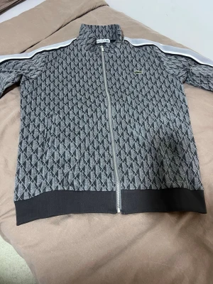 Mönstrad ziptröja från Lacoste - Snygg ziptröja från Lacoste i svart och grått med grafiskt diamantmönster. Tröjan har hög krage, vita ränder längs ärmarna och den klassiska gröna krokodilloggan på bröstet. Ribbstickade muddar och nederkant ger en sportig look.