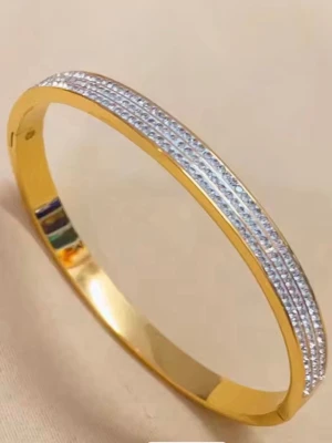 Guldigt armband med gnistrande stenar - Snyggt armband i guldfärgad metall med tre rader små, gnistrande stenar på ovansidan. Armbandet har en stilren och elegant design som verkligen fångar ljuset. Perfekt för dig som gillar accessoarer som sticker ut och ger en lyxig känsla.