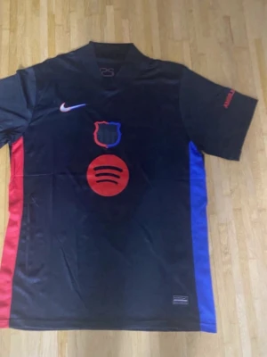 Svart FC Barcelona matchtröja Nike - Snygg svart FC Barcelona matchtröja från Nike med röd och blå detalj på sidorna. Tryck med Spotify-logga och klubbmärke framtill, UNHCR-logga bak. Korta ärmar och sportig passform, perfekt för fotboll eller träning.