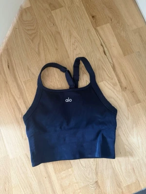 Mörkblå ribbad sport-bh från Alo - Snygg mörkblå sport-bh från Alo med ribbad struktur och justerbara axelband. Tillverkad i ett stretchigt material av nylon och elastan som ger bra stöd och komfort under träningen. Liten vit Alo-logga framtill.