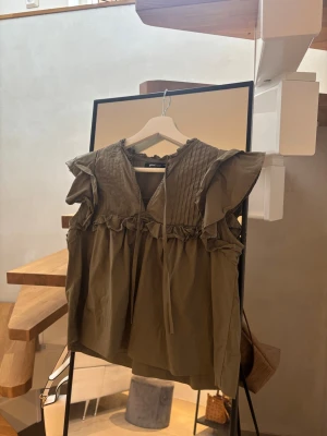 Olivgrön volangblus - Snygg olivgrön blus med volangdetaljer och rysch längs axlar och bröst. Blusen har v-ringning med knytband och dekorativa sömmar framtill. Tillverkad i ett mjukt och avslappnat material. 