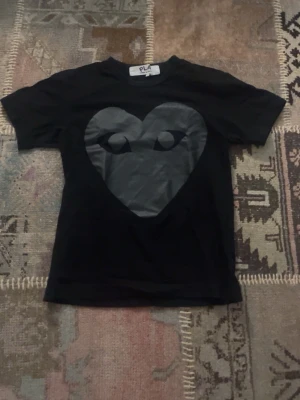 Comme des Garçon t-shirt  - Svart t-shirt från CDG med stort hjärtmotiv i mörkgrått på både fram- och baksidan. På framsidan har hjärtat ögon som ger en cool och unik look. Klassisk rund hals och korta ärmar. Perfekt för dig som gillar statement-plagg.