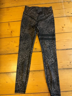 Leopardmönstrade leggings med svarta detaljer - Säljer ett par snygga leopardmönstrade leggings i svart och grått. De har hög midja och två svarta band runt ena låret för en cool look. Perfekta för dig som vill sticka ut lite extra! Stretchigt material som sitter skönt. Och det är från stronger