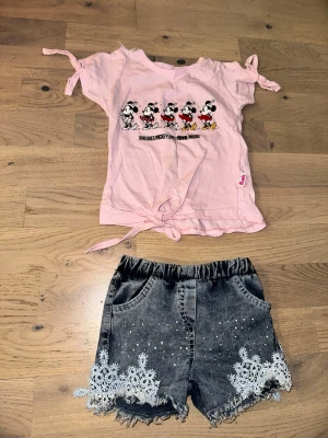 Svarta grå  shorts med spets och pärlor och rosa Mickey mouse t shirt - Säljer ett par svarta shorts med snygg spetsdetalj och pärlor längst ner på benen. Shortsen har fransig kant, två framfickor och resår i midjan för skön passform. Perfekta till sommaren för en trendig look.