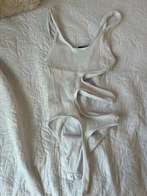 Vit ribbad body med cut-outs - Trendig vit body tröja i ribbat material med flera cut-outs på sidan för en edgy look. Smala axelband och knäppning i grenen. Perfekt för dig som vill sticka ut på stranden eller vid poolen.