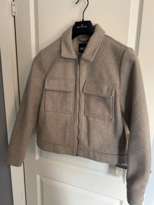 Vårjacka - Stilren beige vårjacka från Vero Moda med två stora fickor och dragkedja framtill. Den har en rak passform, klassisk krage och är tillverkad i ett mjukt, ull-liknande material dock är den nopprig om man tittar närmare på den vilket man ser på den tredje bilden.