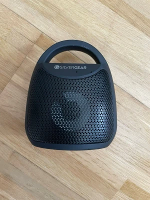 Silvergear bärbar högtalare svart - Bärbar högtalare från Silvergear i svart plast med rundad form och praktiskt handtag. Högtalaren har ett perforerat mönster på framsidan och gummifötter på undersidan för stabilitet. Perfekt för att ta med sig och spela musik var du än är.