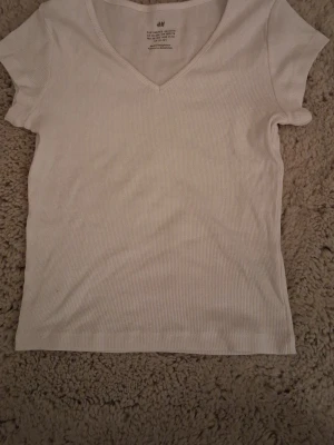 Vit ribbad t-shirt från H&M - Klassisk vit ribbad t-shirt från H&M med v-ringning och korta ärmar. Tillverkad i mjuk bomull som känns skön mot huden. Enkel och stilren modell som passar till det mesta.