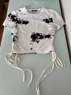 Vit och svart ribbad topp med snörning - Trendig ribbad topp i vitt med svart tie-dye mönster. Kortärmad modell med snörning i sidorna för justerbar passform och croppad look. Perfekt för en cool och avslappnad stil.