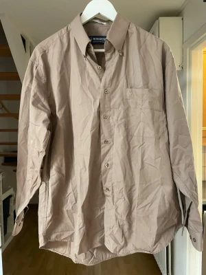 Beige skjorta från Monarch i bomull - Säljer en klassisk beige skjorta från Monarch i 100% bomull. Skjortan har lång ärm, bröstficka och knappar framtill. Perfekt för dig som gillar enkel och stilren design. 