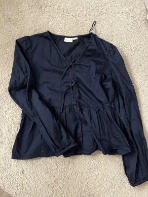 Mörkblå blus med knyt från Gina Tricot - Snygg mörkblå blus från Gina Tricot med v-ringning och dekorativa knytband framtill. Blusen har långa ärmar och en lös, flowy passform med rynkade detaljer under bysten. Perfekt att styla med jeans eller kjol för en trendig look.andvänd edanst 1 gång 