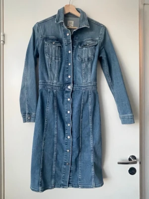 Jeansklänning från Lee med knappar - Säljer en klassisk blå jeansklänning från Lee med långa ärmar och knappar hela vägen framtill. Klänningen har två bröstfickor med lock och markerad midja för en snygg siluett. Perfekt för dig som gillar denim och tidlös stil.