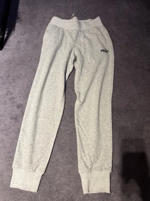 Grå mjukisbyxor från Puma - Snygga ljusgrå mjukisbyxor från Puma med resår i midjan och muddar vid bensluten. Byxorna har en klassisk passform och Puma-logga på vänster lår. Tillverkade i mjukt bomullsmaterial för maximal komfort.