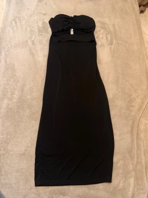 Svart axelbandslös långklänning - Stilren svart långklänning med axelbandslös design och lätt rynkad detalj vid bysten. Klänningen har en smickrande passform och är tillverkad i ett mjukt, stretchigt material som ger en elegant siluett. Perfekt för dig som vill ha en klassisk look.