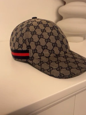 Gucci keps med GG-mönster - Snygg keps från Gucci i beige med klassiskt GG-monogram över hela kepsen. På sidan finns en ikonisk röd och marinblå rand. Kepsen har böjd skärm och är tillverkad i ett slitstarkt tygmaterial. Perfekt för dig som vill ha en trendig och exklusiv accessoar.
