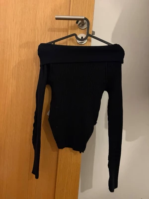 Svart ribbad offshoulder topp - Trendig svart ribbad offshoulder topp med lång ärm. Sitter tight och har en bred kant upptill som ger en snygg siluett. Perfekt att styla med höga jeans eller kjol för en cool look.