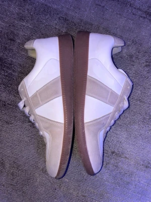 Vita och beige sneakers från Maison Margiela - Snygga sneakers från Maison Margiela i vitt och beige läder och mocka. Skorna har klassisk gummisula i brunt, vita snören och en stilren design med paneler i mocka på sidorna. Perfekta för dig som gillar minimalistisk och trendig stil.