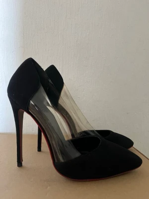 Svarta pumps med transparent detalj 11cm - Snygga svarta pumps med spetsig tå och hög smal klack. Skorna har en cool transparent plastdetalj på sidorna och röd sula som ger extra edge. Perfekta för dig som vill sticka ut lite extra. Materialet är en mix av mocka och plast.
