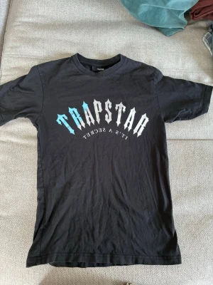 Svart Trapstar t-shirt med tryck - Säljer en svart t-shirt från Trapstar med stort grafiskt tryck i vitt och blått på bröstet där det står 'Trapstar It's a Secret'. Klassisk rund hals och korta ärmar. Perfekt för dig som gillar streetwear. Priset är inte hugget i sten.