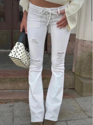 Vita bootcut jeans med slitningar - Snygga vita bootcut jeans med trendiga slitningar på låren och knäna. Hög midja med snörning framtill ger en unik look. Perfekta för dig som vill sticka ut och gillar en modern stil.