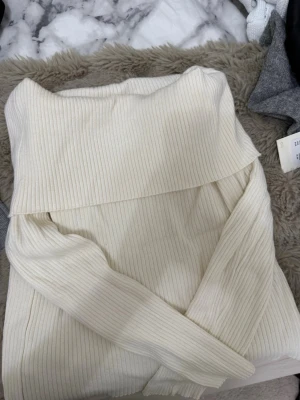 Off shoulder stickad tröja cremevit - Trendig off shoulder stickad tröja i cremevitt med ribbad struktur. Långa ärmar och bred krage som viks ner över axlarna. Perfekt för dig som vill ha en stilren och modern look.