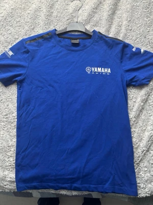 Blå Yamaha Racing t-shirt - Snygg blå Yamaha Racing t-shirt med vita tryck på bröstet och ärmarna. T-shirten har klassisk passform, rund hals och korta ärmar. Materialet är mjuk bomull och trycken inkluderar Yamaha-loggan samt Yamalube på ena ärmen.