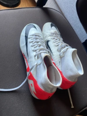 Nike vita och röda fotbollsskor - Säljer ett par Nike fotbollsskor i vitt med röda och svarta detaljer. Skorna har hög krage för extra stöd, snörning och dobbar för bra grepp på planen. Tillverkade i syntetmaterial som ger lätt känsla och bra bollkontroll. Perfekta för dig som vill sticka ut på planen.