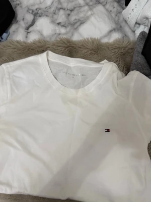 Vit t-shirt från Tommy Hilfiger - Klassisk vit t-shirt från Tommy Hilfiger med rund halsringning och liten broderad logga på bröstet. Tillverkad i mjuk bomull och har en enkel, stilren design som passar till det mesta.