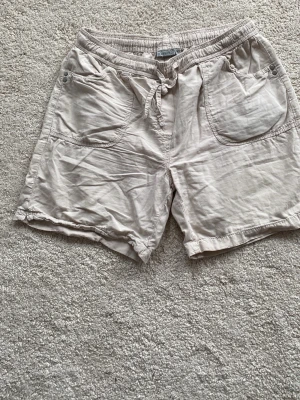 Beige shorts med snörning från Cellbes - Säljer ett par beige shorts från Cellbes med elastisk midja och snörning. Shortsen har stora fickor framtill med dekorativa nitar och är tillverkade i mjuk bomull. Perfekta för varma dagar och enkel att matcha med olika toppar.