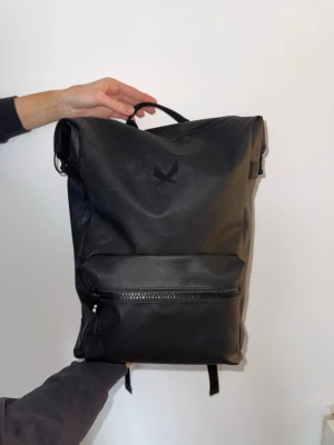 Svart ryggsäck från SOC - Snygg svart ryggsäck från SOC i stilren design. Tillverkad i vattenavvisande material med en ficka framtill och dragkedja. Justerbara axelband och bärhandtag upptill. Köpt från Stadium. Har flera vita fläckar på sig men i övrigt bra skick.