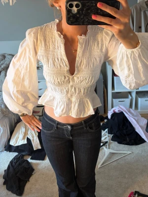Vit smockad blus med volang - Trendig vit blus med smockad överdel, djup v-ringning och volang nedtill. Blusen har puffiga långärmar med resår vid ärmslut och är tillverkad i ett lätt bomullsliknande material. Perfekt för en chic och avslappnad look.