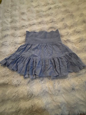 Blå volangkjol med smock - Jättegullig blå kjol med bred smock i midjan och volangkant. Kjolen har små rosa blommor och är gjord i ett fint viskos tyg. Perfekt för sommarens alla tillfällen och ger en avslappnad men ändå trendig look.