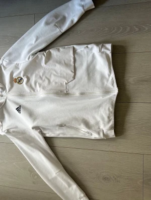Vit Real Madrid hoodie från Adidas - Snygg vit hoodie från Adidas med Real Madrid-logga på bröstet och dragkedja framtill. Tröjan har långa ärmar, två sidofickor med dragkedja och Adidas-logga. Tillverkad i mjukt material.
