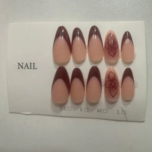 Brun röda  press-on naglar med blommor - Snygga press-on naglar i mandelform med brun fransk tipp och två naglar med blommönster. Färgen är nude i botten och mörkbrun på tippen. Perfekt för dig som vill ha en trendig och elegant look. Kommer i flera storlekar.