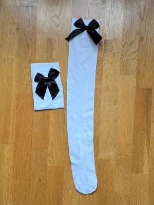 Vita strumpor med svart rosett - Snygga vita strumpor med en stor svart rosett upptill. Tightsen har en klassisk passform och är tillverkade i ett stretchigt material som sitter bekvämt på benen. Perfekta för att ge din outfit en söt och trendig touch.