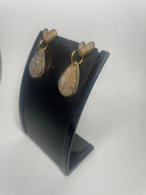 Guldiga örhängen med hjärta och droppe - Unika örhängen med hjärtformad topp och droppformad hängande del i guldiga och beige toner. Örhängena har en marmorerad effekt och är tillverkade i metall och resin för en modern look. Perfekta för dig som vill sticka ut med snygga detaljer.