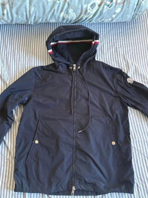 Mörkblå vindjacka från Moncler - Snygg mörkblå vindjacka från Moncler med huva och dragkedja framtill. Jackan har två fickor med tryckknappar och Moncler-logga på ärmen. Detaljer med röd och vit rand på huvan ger en sportig touch. Tillverkad i lätt, vattenavvisande material. dragkedjan funkar perfekt, bara lite kosmetisk rost.