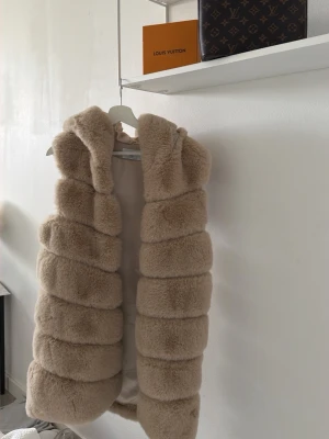 Beige fluffig väst - Supersnygg beige pälsväst i fuskpäls med breda, horisontella paneler och stor, mysig krage. Västen är ärmlös och har en lyxig känsla som passar perfekt till lager-på-lager outfits. Ingen synlig branding.