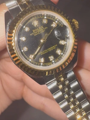 Rolx Datejust tvåtonad klocka - Snygg Rolx Datejust 28 mm med svart urtavla och guldfärgade detaljer. Klockan har en tvåtonad länk i rostfritt stål och guld, räfflad guldfärgad bezel och indexmarkeringar med gnistrande stenar. Klassisk och lyxig design. Oanvänd. 