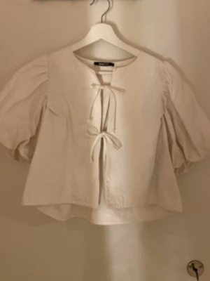 Vit blus med puffärmar och knyt - Söt vit blus med puffiga trekvartsärmar och två knytband framtill. Blusen har en avslappnad passform och rund halsringning. Perfekt att styla med jeans eller kjol för en trendig look.