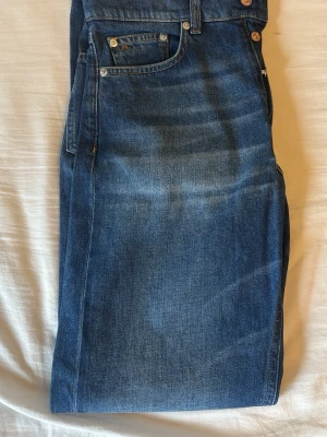 Blå raka jeans i klassisk modell från J.LINDBERG - Säljer ett par blå jeans i klassisk rak modell med fem fickor och normalhög midja. Jeansen har en snygg tvätt med subtila slitningar och är tillverkade i kraftigt denimtyg. Perfekta för dig som gillar tidlös stil och enkelhet.