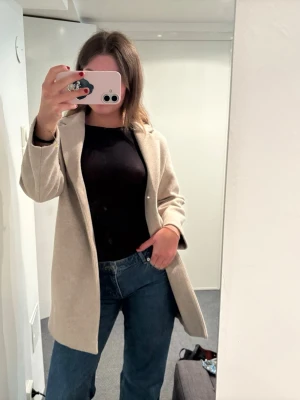 Beige rak kappa i ullmix - Stilren beige kappa med rak passform och enkel design. Kappan har diskreta fickor i sidorna och är tillverkad i en mjuk ullmix som passar perfekt till höst och vinter. Inga synliga knappar framtill för en clean look.