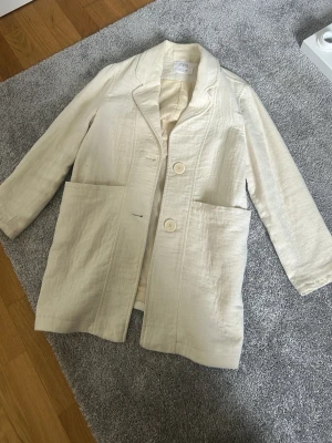 Krämvit kavaj från Zara Kids - Stilren krämvit kavaj från Zara Kids med två stora framfickor och två knappar framtill. Tillverkad i ett lätt linneliknande material som ger en avslappnad men ändå snygg look. Perfekt för att lyfta vilken outfit som helst.