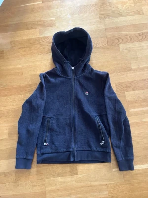 Mörkblå hoodie från Fila - Snygg mörkblå hoodie från Fila med dragkedja framtill och två fickor med dragkedja. Liten Fila-logga på bröstet och coolt band med rödvita ränder på ryggen. Tillverkad i mjukt, strukturerat material och har ribbade muddar vid ärmar och nederkant.