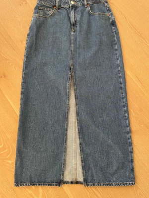 Lång jeanskjol Never Denim Bik Bok  - Klassisk lång jeanskjol från Bik Bok i blå denim. Kjolen har hög midja, femficksmodell och en snygg slits framtill för extra rörelsefrihet. Perfekt till sneakers eller boots för en avslappnad look.