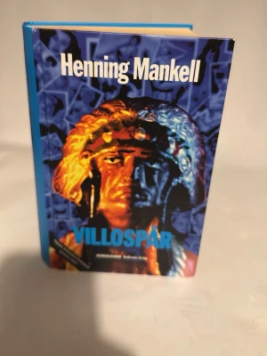Villosprår av Henning Mankell - En spännande kriminalroman av Henning Mankell med hård pärm i blått och ett färgstarkt omslag med motiv i orange, blått och svart. Boken är på svenska och tillhör Wallander-serien. Perfekt för dig som gillar deckare och nordisk spänning.