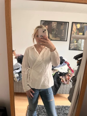 Vit omlottblus med knytband - Säljer en stilren vit omlottblus med knytband i sidan. Blusen har lång ärm och en klassisk krage. Passar perfekt till både jeans och kjol för en trendig look.