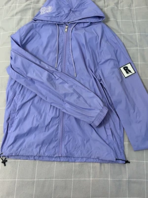 Ljuslila vindjacka med huva - Snygg ljuslila vindjacka med huva och dragkedja framtill. Jackan har ett vitt patch med svart motiv på ena ärmen och justerbar dragsko nedtill. Perfekt för blåsiga dagar och enkel att matcha till olika outfits.